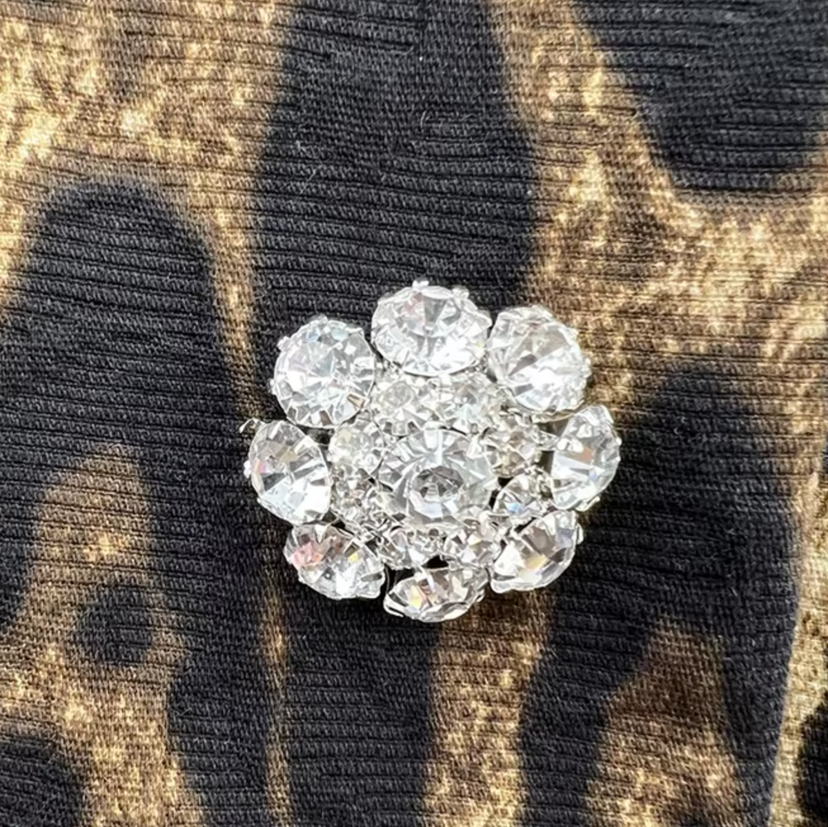A round cluster brooch made of clear rhinestones in a silver-tone setting, displayed on a black and gold patterned fabric background.