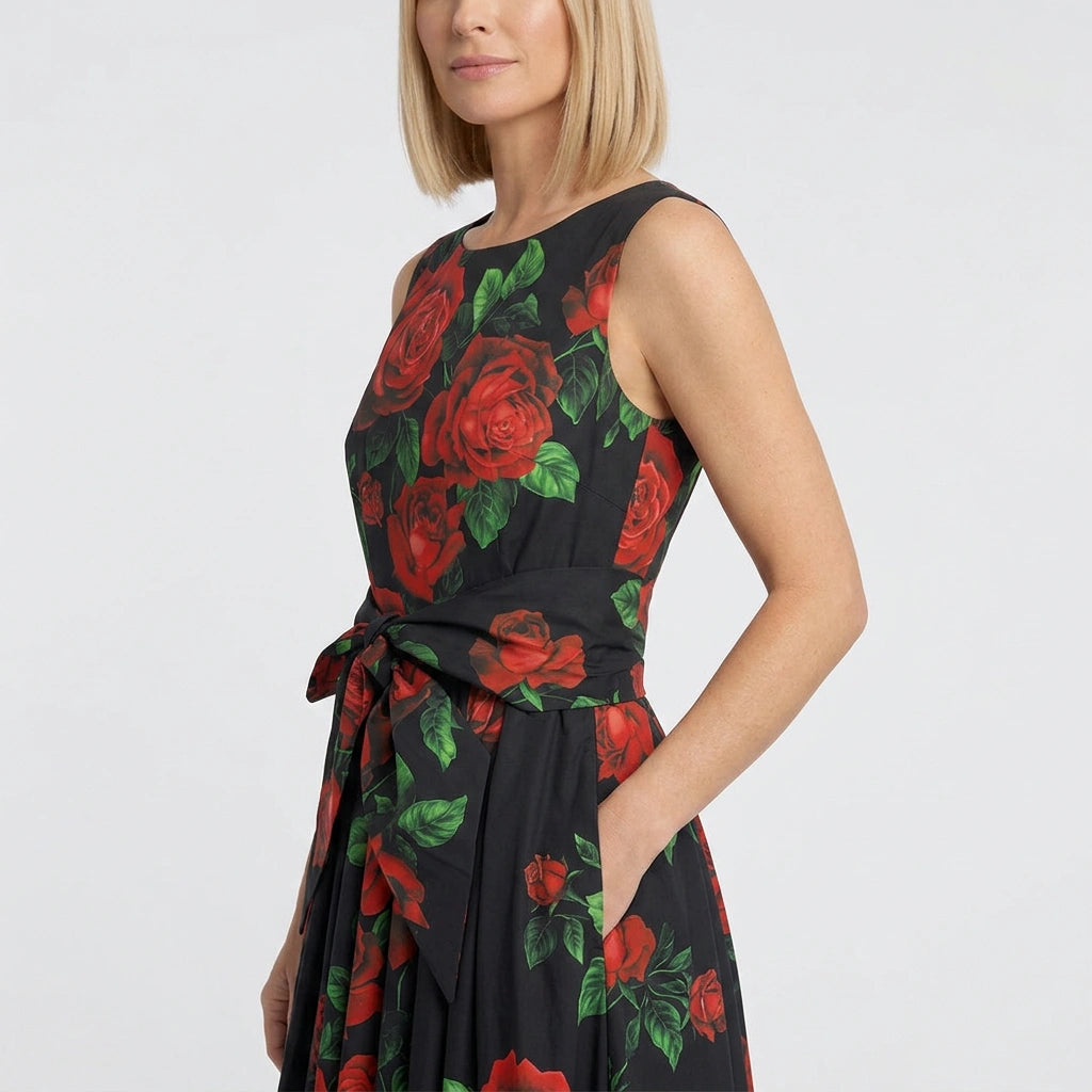 A woman wearing a sleeveless black dress with a red rose and green leaf floral print, tied at the waist, shown against a plain light gray studio background.