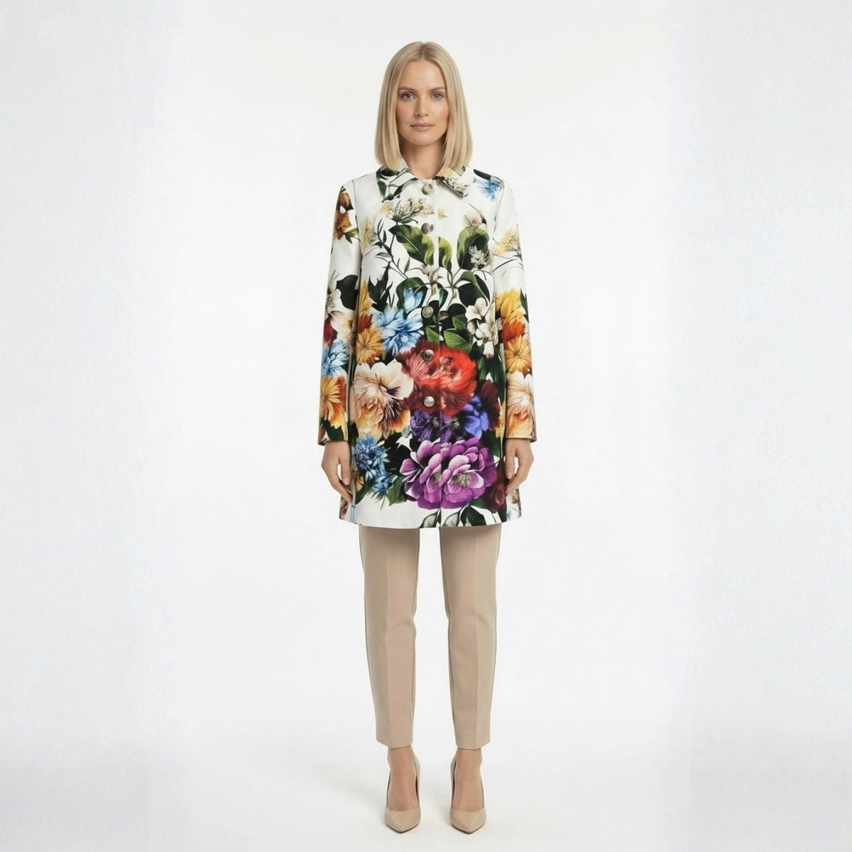 A woman stands against a plain white studio background wearing a long-sleeve white brocade coad with a large multicolor floral print, paired with beige cropped pants and nude heels.