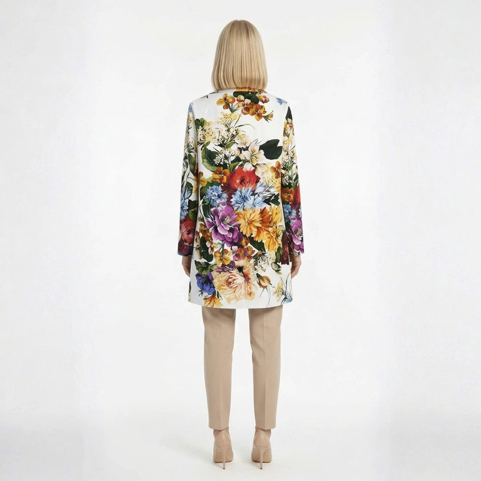 A woman stands against a plain white studio background wearing a long-sleeve white brocade coad with a large multicolor floral print, paired with beige cropped pants and nude heels.