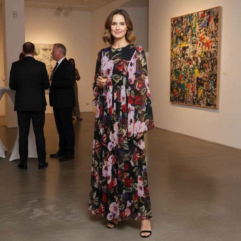 A woman wearing a long-sleeved black maxi dress with a vibrant pink, red, and green floral print stands in an art gallery. In the background, several men in black suits are mingling near a white reception table, and colorful abstract paintings are hanging on the gallery walls.