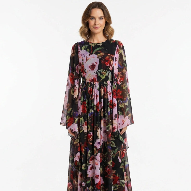 A woman modeling a long-sleeved maxi dress with a black base and a vibrant pink, red, and green floral print, set against a plain white studio background.