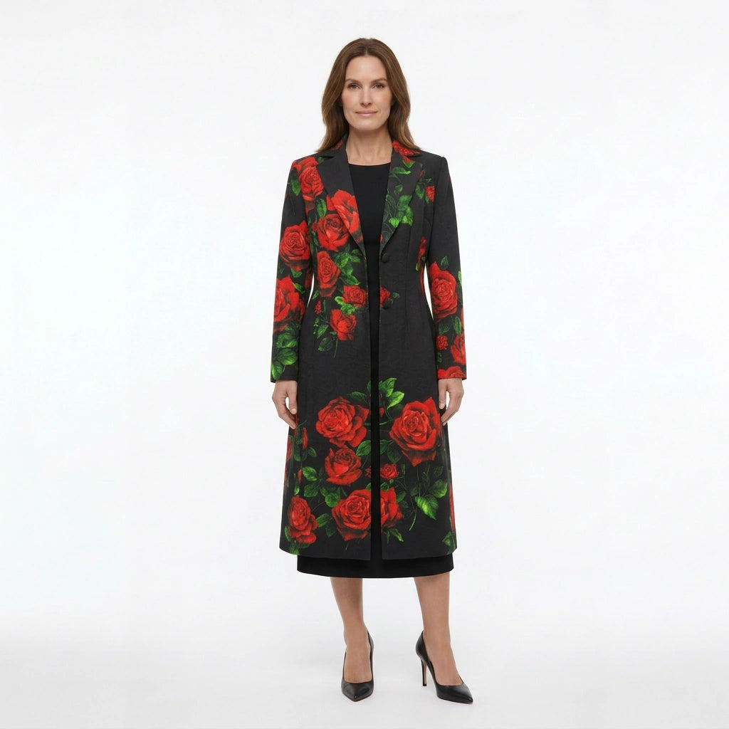 A woman modeling a long black coat with a vibrant red rose and green leaf pattern, paired with a black dress and black pointed-toe heels, against a plain white studio background.