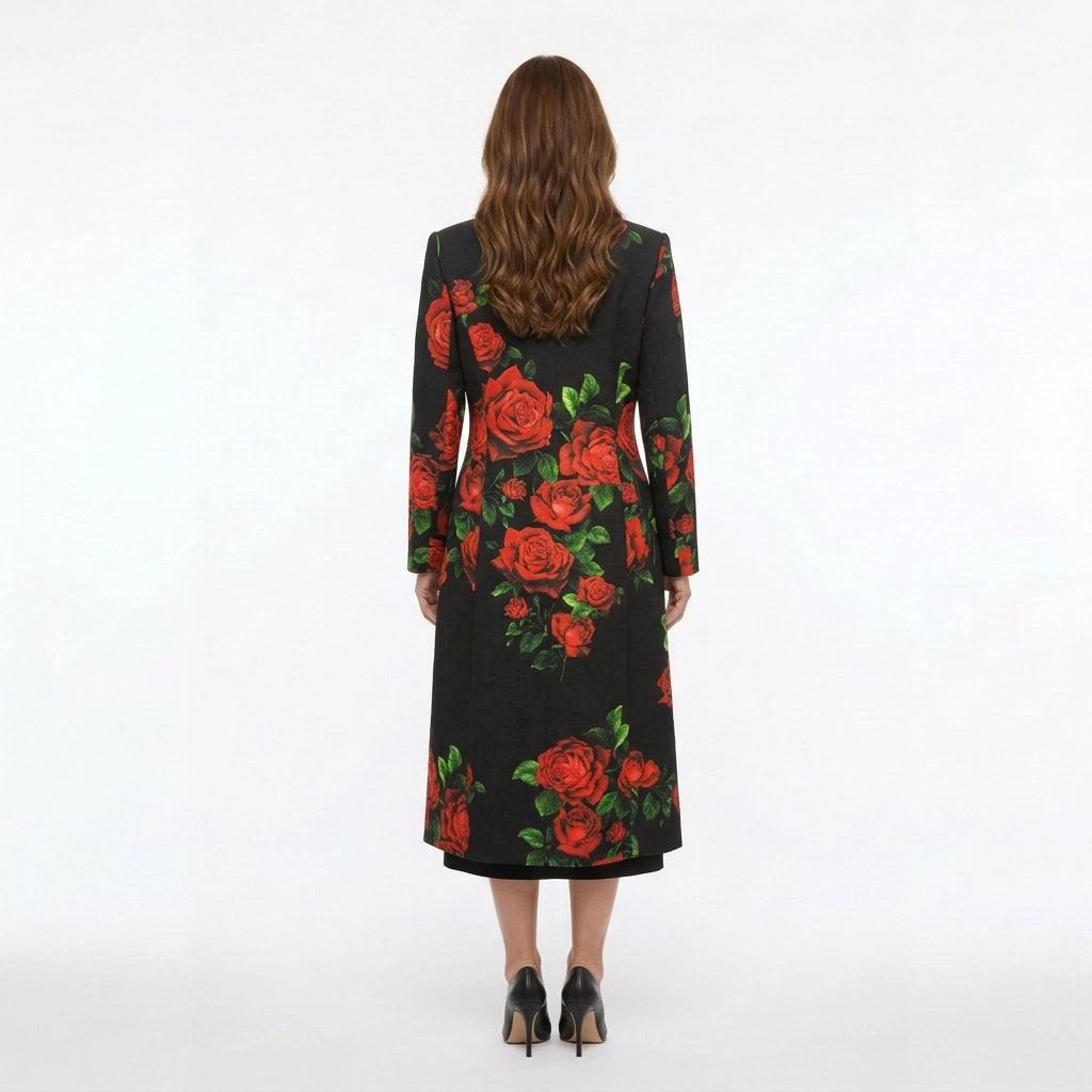 A woman viewed from behind wearing a long black coat with a bold red rose and green leaf floral print, paired with black heels, against a plain white studio background.