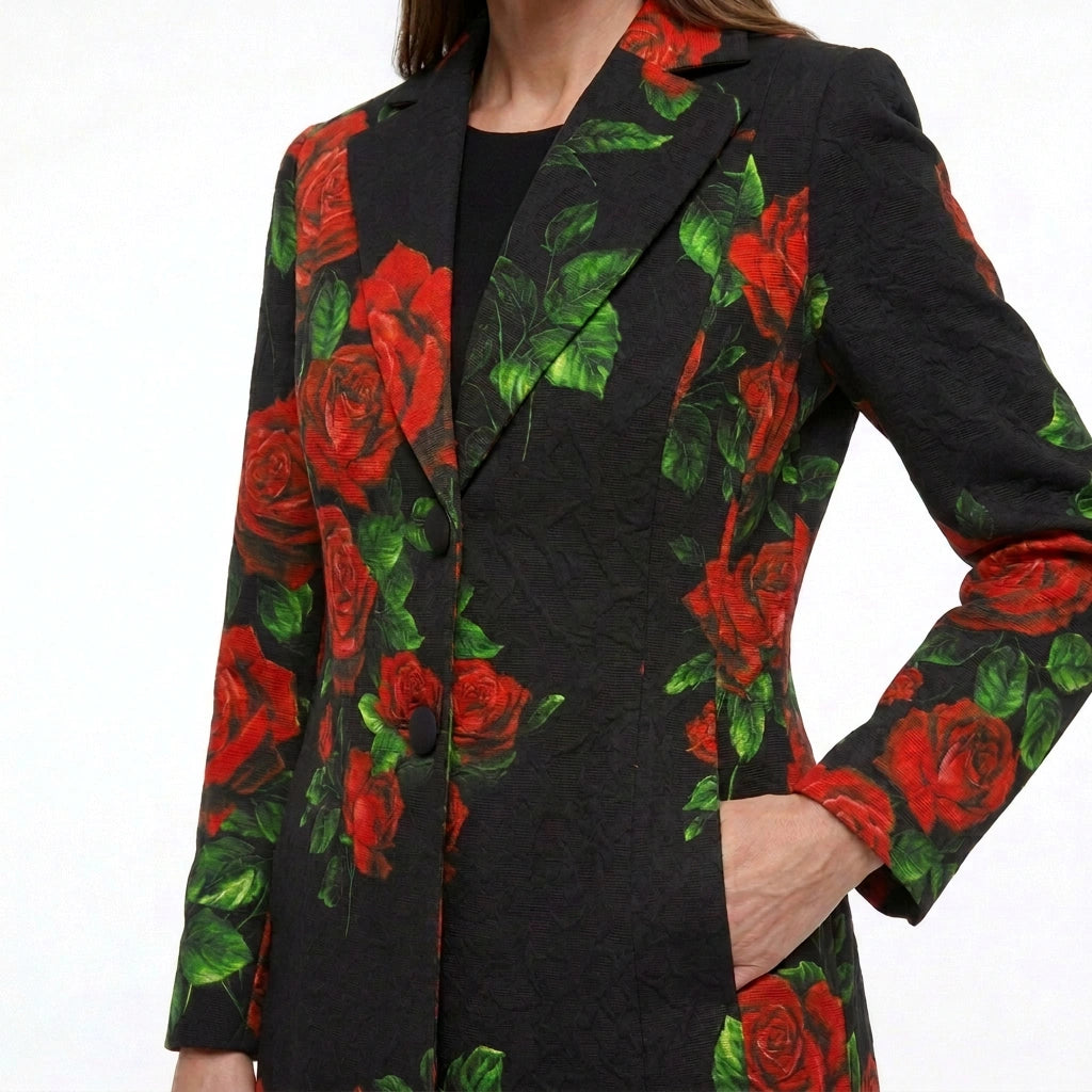 A close-up of a model wearing a black textured blazer with a vibrant red rose and green leaf pattern, paired with a black top against a plain white background.